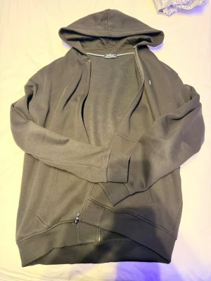 Svart hoodie från Polo Ralph Lauren XL-L - Klassisk svart hoodie från Polo Ralph Lauren i storlek XL-L. Tröjan har huva, dragkedja framtill och fickor. Tillverkad i mjuk bomull med det ikoniska broderade logotypen på bröstet. Perfekt för chill dagar eller när du vill ha en clean streetstyle.