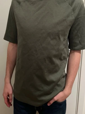 Grön t-shirt från BOSS, storlek M - Snygg grön t-shirt från BOSS i storlek medium. Modellen har korta ärmar och en clean, rak passform som ger en chill vibe. Materialet är mjukt och känns som bomull, perfekt för dig som gillar stilrena och enkla plagg. Inga mönster eller detaljer, bara klassisk look.