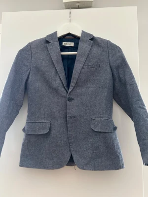 Grå/blå jeans kavaj från H&M med mockadetalj - Snygg jeans kavaj från H&M med klassisk passform och två framfickor. Kavajen har bruna mockalappar på armbågarna och är helfodrad med mörkblått tyg. Perfekt för dig som vill ha en stilren look med lite extra detaljer.