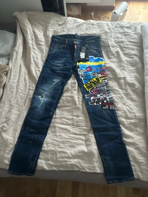 Dsquared2 jeans med patchwork och tryck - Helt oanvända Dsquared2 jeans i storlek 46 med patchwork, färgstarka tryck och broderade detaljer på ena benet. Jeansen har raka ben, slitningar och färgstänk för en edgy look. Säljer då jag ej har användning av dessa. Dessa är äkta. 