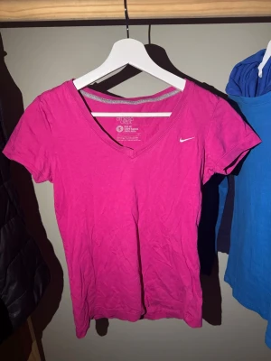 Rosa Nike t-shirt med v-ringning - Säljer en snygg rosa t-shirt från Nike med v-ringad hals och liten logga på bröstet. Modellen är slim fit och har korta ärmar. Perfekt för dig som gillar sportig stil och vill ha något bekvämt i bomull. Vidareförsäljning❤️
