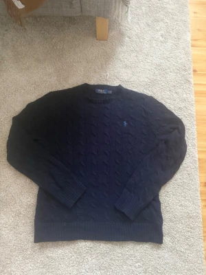 Marinblå stickad tröja Polo Ralph Lauren - Säljer en marinblå stickad tröja från Polo Ralph Lauren i storlek M. Tröjan har klassiskt kabelstickat mönster, rund hals och långärmade ribbade muddar. 