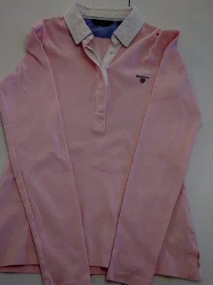 Ljusrosa henley från Gant XS - Säljer en ljusrosa henley från Gant i storlek XS. Skjortan har vit krage, långa ärmar och knappar vid halsen. Klassisk och stilren design med Gant-logga broderad på bröstet. Perfekt för dig som gillar preppy stil.