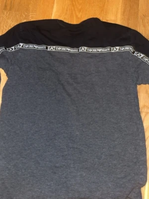 Grå EA7 Emporio Armani t-shirt - Säljer en grå t-shirt från EA7 Emporio Armani med svart axelparti och vita logoband längs axlarna. Stort EA7-tryck på ryggen och klassisk rund hals. Skön och stilren modell i mjukt bomullsmaterial.