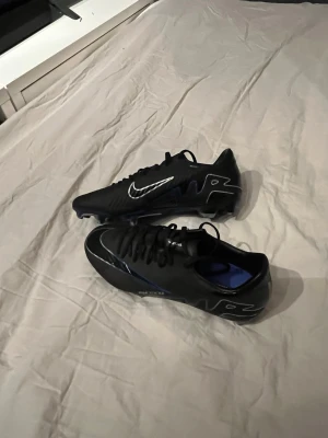 Nike svarta fotbollsskor med blå sula - Säljer ett par svarta Nike fotbollsskor med blå yttersula och metall-dobbar. Skorna har snörning och en snygg silverfärgad swoosh på sidan och framtill. Ovandelen är gjord i syntetmaterial och insidan är blå. Perfekt för dig som vill ha grymt grepp på planen.