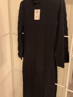 Svart långärmad klänning från JDY - Säljer en svart långklänning från JDY med långärmade ärmar och en unik, skrynklig textur. Klänningen har rund halsringning och är enkel men ändå trendig. Perfekt för dig som gillar minimalistisk stil och vill ha något som sticker ut lite med sin struktur. Helt oanvänd med prislappen på. 