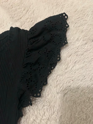 Svart blus med volangärmar från Zara - Säljer en svart blus från Zara i storlek S. Toppen har ribbad kropp och korta, breda volangärmar med broderade detaljer och hålmönster. Rund halsringning och figurnära passform. Perfekt för dig som gillar unika ärmdetaljer och stilren look.