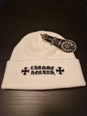 Vit Chrome Hearts mössa - Säljer en vit mössa från Chrome Hearts med broderad logga och svarta korsdetaljer framtill. Mössan har uppvikt kant och är tillverkad i mjukt stickat material som känns skönt mot huvudet. Perfekt för att lyfta din streetwear-look.