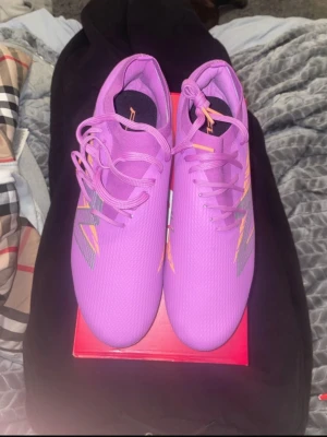 New Balance V8 PRO  - Säljer ett par lila New Balance fotbollsskor med orange och silver detaljer. Skorna har snörning och en platt sula med orange dobbar för gräsplan. Ovandelen är tillverkad i syntetmaterial med ett mönstrat tryck på sidan. Perfekt för dig som vill sticka ut på planen.