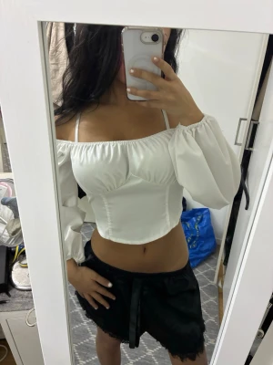Vit offshoulder korsett-topp - Säljer en vit korsett-topp med offshoulder och puffiga långärmar. Toppen har smockad rygg, croppad passform och tunna axelband. Materialet är mjukt och har en lätt glans. 