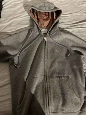Grå zip-hoodie från Burberry - Grå hoodiejacka från Burberry med dragkedja, huva med snörning och klassiskt rutigt foder. Jackan har broderad logga på bröstet och är tillverkad i mjuk bomullsmix. Perfekt för dig som gillar stilren och bekväm streetwear.
