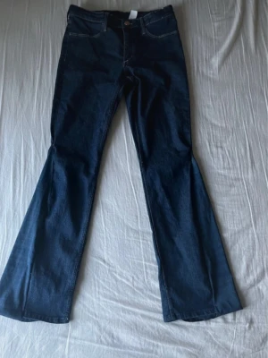 Mörkblå bootcut jeans - Snygga mörkblå jeans med bootcut-snitt och klassisk femficksdesign. Jeansen har en markerad midja och är tillverkade i ett slitstarkt denimtyg. Passar perfekt om du gillar retrovibbar och vill ha ett par jeans med lite extra vidd nedtill. 