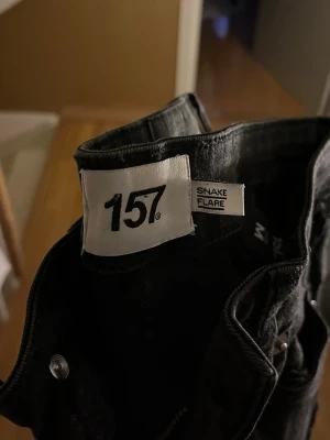 Svarta Snake Flare jeans från 157 - Svarta jeans från 157 i modellen Snake Flare. Byxorna har klassisk femficksdesign och en snygg flare-silhuett som ger en cool vibe. Materialet är jeans och färgen är helsvart, perfekt för dig som gillar stilrena och trendiga byxor.