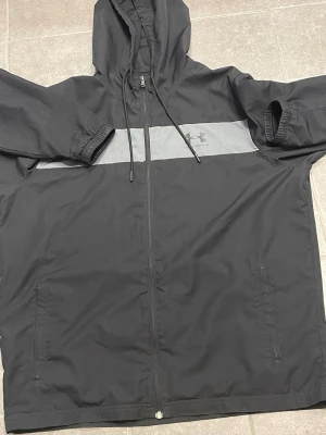 Svart vindjacka från Under Armour - Svart vindjacka från Under Armour med grått band över bröstet och logga. Jackan har huva med snörning, hel dragkedja och två sidofickor. Materialet är lätt och syntetiskt, perfekt för blåsiga dagar. Passformen är normal och ärmarna är långärmade med resår vid mudden.