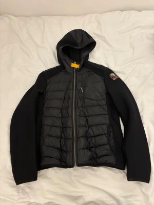 Svart pufferjacka från Parajumpers - Svart pufferjacka från Parajumpers med huva och coolt gult spänne vid halsen. Jackan har quiltad framsida och rygg, dragkedjor på fickor och bröst, samt patch med logga på ärmen. Materialmix av polyester och syntet, perfekt för höst och vinter.