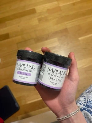 Saviland Builder Nail Gel Clear & Milky White - Två burkar builder nail gel från Saviland, en i färgen Clear och en i Milky White. Gelen används för att bygga och förlänga naglar, ger en snygg och naturlig finish. Burkarna är svarta med lila och vita etiketter, innehåller 100g vardera.