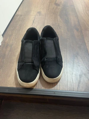 Axel arigato skor - Svarta slip-on sneakers med vit platt sula och rund tå. Ovandelen är gjord i mocka och har ett brett elastiskt band över foten för enkel påtagning. Skorna har en stilren och enkel design som passar till många olika outfits köpt dom för 500kr nypris säljer för 327kr. Då skor passa ej mig längre 