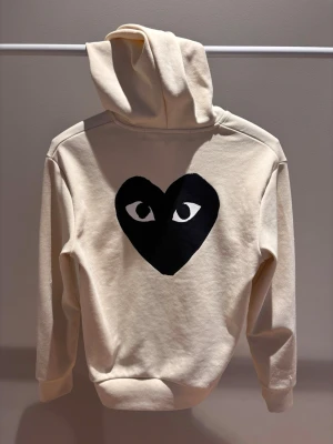Comme des garcons Hoodie - Snygg hoodie i cream/beige färg, storlek Small. Tröjan har en bra passform och passar personer mellan ca 160–175 cm. Tillverkad i mjukt och bekvämt material med hel dragkedja, huva med dragsnören och praktiska framfickor. Stilren design med svart hjärtlogga på bröstet och större matchande tryck på ryggen. I bra skick med endast lätt användning. Enkel att matcha och perfekt för både vardag och fritid.