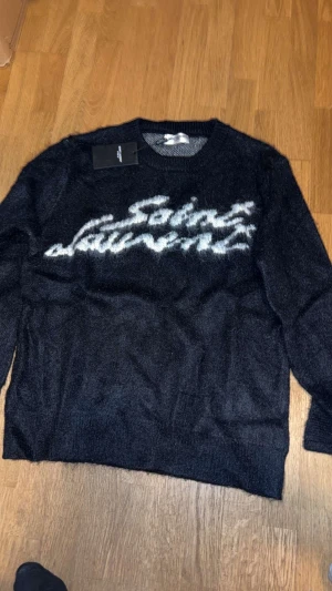 Stickad Saint Laurent tröja - Säljer en svart stickad tröja från Saint Laurent i storlek m med vit, suddig logga över bröstet. Riktigt fin hör av er vid funderingar priset är inte ristat i sten 