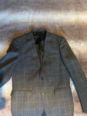 Rutig kavaj från Bläck i mörkgrå - Snygg mörkgrå kavaj från Bläck med rutigt mönster och två knappar framtill. Kavajen har klassisk krage, innerfoder i svart och raka fickor. Perfekt för dig som gillar stilrena och moderna plagg med skandinavisk touch.