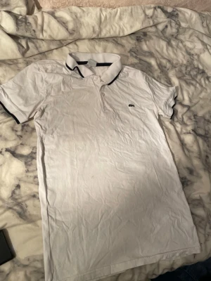 Vit piké från Lacoste, storlek L - Säljer en klassisk vit pikétröja från Lacoste i storlek L. Tröjan har korta ärmar, krage med svarta detaljer och knäppning framtill. Ikoniska Lacoste-loggan med krokodil är broderad på bröstet. Perfekt för en clean och sportig look.