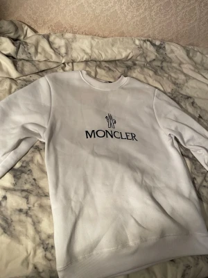Vit sweatshirt från Moncler - Säljer en stilren vit sweatshirt från Moncler med svart logotyp tryckt på bröstet. Tröjan har rund hals och långa ärmar, tillverkad i mjuk bomull som är skön mot huden. Perfekt för dig som gillar exklusiva märken och enkel design.