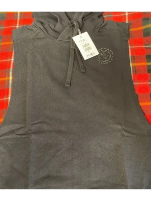 Gymshark svart ärmlös linnetopp med huva - Svart ärmlös linnetopp från Gymshark med huva och snörning framtill. Toppen har en sportig look och är perfekt för träning. Materialet känns mjukt och lätt mot huden. Diskret logga på bröstet. Snygg och enkel att matcha med träningskläder.