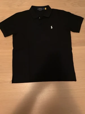 Svart piké från Polo Ralph Lauren - Säljer en svart pikétröja från Polo Ralph Lauren i modellen Custom Slim Fit. Tröjan har korta ärmar, klassisk krage och knappslå framtill. På bröstet finns den ikoniska broderade loggan i vitt. Perfekt till jeans eller shorts!