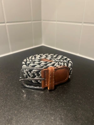 Svart och grå flätad väst - Snygg flätad väst i svart och grått med bruna skinn-detaljer vid spännet och ändarna. Västen har ett randigt mönster och är tillverkad i syntetmaterial med inslag av skinn. Perfekt för dig som vill ha en cool och stilren look.