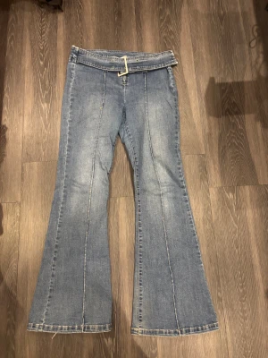 Blå bootcut jeans med bälte - Snygga blå bootcut jeans med markerade sömmar längs benen och ett brett jeansbälte med metallspänne. Jeansen har klassisk dragkedja och är tillverkade i mjukt denimtyg. Perfekt för dig som gillar retrovibbar och vill sticka ut lite extra.