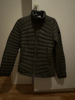 Mörkgrön pufferjacka från Decathlon XS - Säljer en mörkgrön pufferjacka från Decathlon i storlek XS. Jackan har quiltad design, hög krage och två fickor med dragkedja. Perfekt för kyliga dagar och har en clean look som funkar till många stilar.