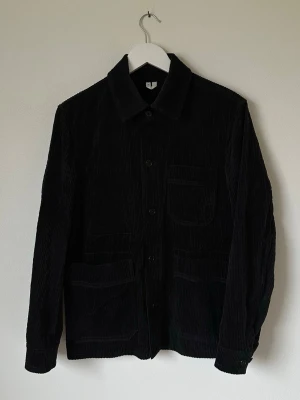Svart manchester overshirt från ARKET - Svart overshirt från ARKET i grov manchester med klassisk krage, knäppning framtill och tre fickor. Perfekt lager-på-lager-plagg med stilren look och långärmad design. Materialet är en bomullsblandning som ger en mjuk och cool känsla. 7Y6