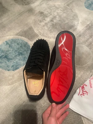 Christian Louboutin svarta sneakers med nitar - Säljer ett par svarta Christian Louboutin sneakers i mocka med coola svarta nitar på tån och klassisk röd sula. Skorna har snörning och rund tå, insida i beige skinn och kommer med dustbag och certifikat. Perfekt för dig som vill sticka ut med lyxig streetstyle.