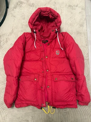 Röd dunjacka från Fjällräven S - Riktigt fet röd dunjacka från Fjällräven med huva, stora fickor med tryckknappar och snörning nertill. Jackan har dragkedja och knappar framtill samt broderad logga på bröstet. Perfekt för kalla dagar när du vill hålla stilen.
