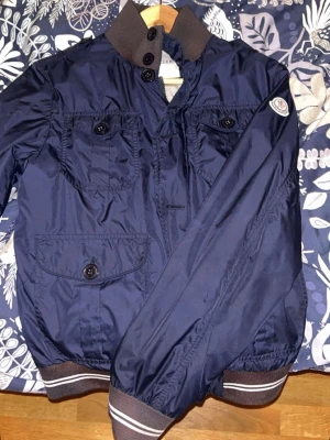 Mörkblå bomberjacka från Moncler - Snygg mörkblå bomberjacka från Moncler med ribbade muddar i brunt och vita ränder. Jackan har två bröstfickor och en ficka nertill, alla med knappar. Klassisk Moncler-logga på ärmen och hög krage med knappstängning. Perfekt för dig som vill ha en stilren jacka.