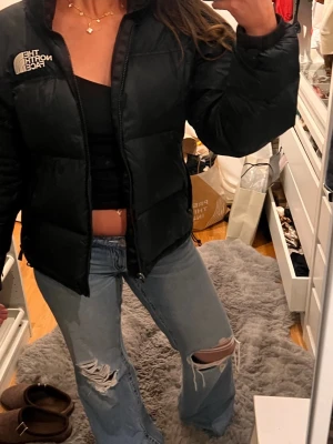 Svart pufferjacka från The North Face - Säljer en svart pufferjacka från The North Face i storlek S. Jackan har en hög krage, synlig dragkedja och logga på bröstet. Den är vadderad och har en loose passform, perfekt för kalla dagar. Materialet är syntet och jackan har praktiska sidofickor.