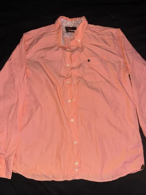 Morris skjorta - Persikofärgad skjorta från Morris med klassisk krage och vita knappar. Insidan av kragen har ett blommigt mönster i rosa, lila och beige. Skjortan har lång ärm och en liten broderad detalj på bröstet. Tillverkad i bomull med Twofold Yarn.