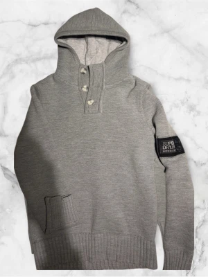 Grå hoodie från Superdry  - Snygg grå hoodie från Superdry med ribbade muddar och huva. Tröjan har tre knappar vid halsen och ett svart Superdry-märke på ärmen. Materialet är stickat och ger en skön känsla. Perfekt för dig som gillar stilren och enkel design. Storlek Medium.
