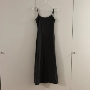Mörkgrå långklänning med smala axelband - Svart långklänning med smala axelband och rundad halsringning. Klänningen har en enkel och stilren design som faller rakt ner och ger en snygg siluett. Materialet ser ut att vara mjukt och följsamt, oanvänd