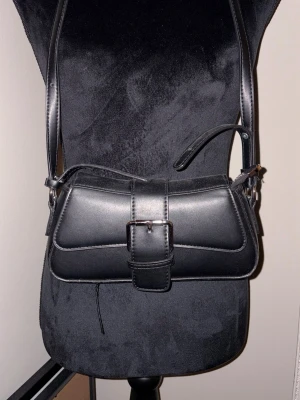 Svart axelväska med spänne - Snygg svart axelväska i skinnimitation med stilrent silverfärgat spänne framtill. Väskan har en klassisk rektangulär form och justerbar axelrem. Perfekt storlek för det viktigaste och passar till många olika outfits.