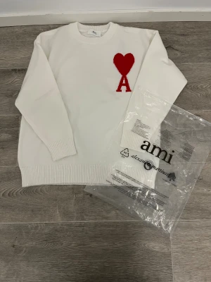 Vit stickad tröja från Ami Paris - Vit stickad tröja från Ami Paris med ett stort rött hjärta och bokstaven A broderat på bröstet. Tröjan har rund hals, ribbade muddar och nederkant samt långärmad passform. Tillverkad i Portugal och har en clean, stilren look som sticker ut med den röda detaljen.