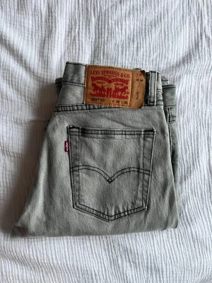 Levi's 501 grå jeans W29 L32 - Säljer ett par klassiska Levi's 501 jeans i grått. Modellen har raka ben, fem fickor och knappgylf. Jeansens material är bomull och de har den ikoniska läderpatchen bak i midjan. Perfekt för dig som gillar tidlös stil och vill ha ett par snygga jeans.