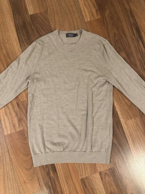 Beige merinoull tröja storlek M - En stilren beige stickad tröja från Dressmann med rund hals och långa ärmar. Tröjan har ribbade muddar vid ärmslut och nederkant, vilket ger en snygg passform. Perfekt för lager-på-lager under hösten och vintern. Bra skick och är storlek Medium.