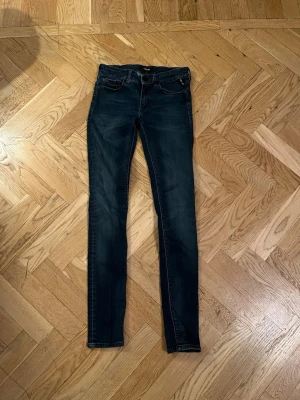 Replay mörkblå slimfit jeans - Snygga mörkblå jeans från Replay med slimfit passform och klassisk femficksdesign. Jeansen har svarta detaljer på knappar och bakficka samt Replay-logga vid fickan. Materialet är stretchigt jeans och midjan är normal. Perfekt för dig som gillar en smal siluett.