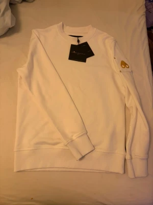 Vit sweatshirt från Moose Knuckles - Vit sweatshirt från Moose Knuckles med guldfärgad metallpatch på ärmen och ribbade muddar vid hals, ärmslut och nederkant. Tröjan har en clean look och är tillverkad i mjuk bomull. Perfekt för dig som gillar stilrena och exklusiva detaljer.