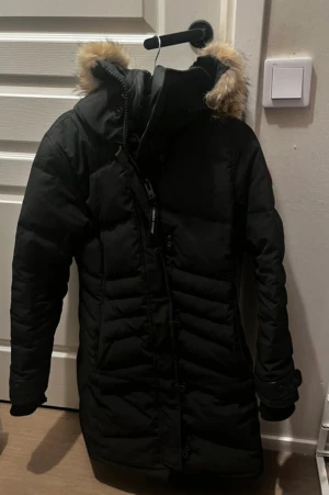 Svart lång dunjacka från Canada Goose - Säljer en svart dunjacka från Canada Goose med lång passform och huva med beige fuskpäls. Jackan har quiltad design, dragkedja och knappar framtill samt ribbade muddar vid ärmslut. Perfekt för kalla vinterdagar och riktigt snygg stil.