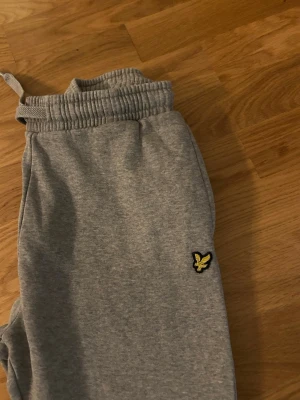 Grå mjukisbyxor Lyle & Scott Junior - Snygga grå mjukisbyxor från Lyle & Scott Junior med klassisk gul fågel-logga på benet. Byxorna har ribbad midja med snörning och muddar vid bensluten. Perfekt för chill eller träning, tillverkade i mjukt bomullsmaterial.
