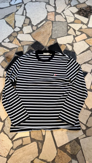Maison Kitsuné randig långärmad tröja - Säljer en svartvit randig långärmad tröja från Maison Kitsuné i storlek M. Tröjan har ett broderat rävlogo på bröstet i blått, vitt och rött. Rund halsringning med svart kant. Tillverkad i mjuk bomull som är skön mot huden.