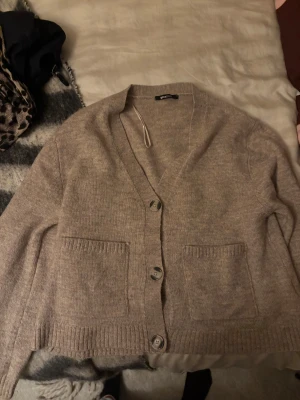 Beige kofta från Gina Tricot - Mysig beige kofta från Gina Tricot i storlek S. Koftan har v-ringning, tre knappar framtill och två praktiska fickor. Ribbstickade detaljer vid ärmslut och nederkant ger en snygg finish. Perfekt att slänga på sig när det är lite kyligt ute.