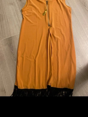 Orange ärmlös klänning Star Park - Snygg orange ärmlös klänning från Star Park med svart fransdetalj längst ner och dragkedja framtill med tofs. Klänningen har rak passform och är tillverkad i ett mjukt syntetmaterial. Perfekt för dig som gillar färg och coola detaljer.